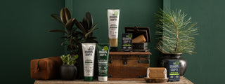 Various Grandpa Soap Co. Pine Tar personal care products, including shampoo, conditioner, body wash, bar soap, a plant, a brown toiletry bag, and a wooden chest, displayed on a dark wooden surface against a green background.