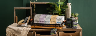 A styled display of Grandpa Soap Co. bar soaps, including Charcoal, Witch Hazel, Epsom Salt, Oatmeal, Thylox, and Pine Tar, arranged on a wooden cabinet with green plants, ceramic vases, and neutral textiles against a dark green wall.