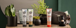 A lifestyle display of Grandpa Soap Co. body care products, including shower creams, body wash, and shower oil, arranged on a wooden vanity with green plants, a round mirror, grooming accessories, and a toiletry bag against a dark green wall.