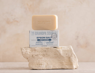 A bar of The Grandpa Soap Co. Epsom Salt soap rests on top of its box, which sits on a textured white stone against a neutral background.