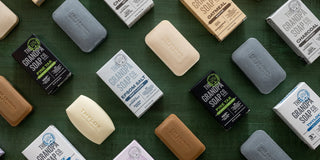 Bars of soap and soap boxes in various colors are neatly arranged on a green surface. The soaps include brands like The Grandpa Soap Co. in different scents and styles, as well as other unbranded soap bars.