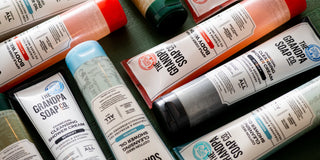 Tubes and bottles of The Grandpa Soap Co. cleansing shower creams and soaps are arranged on a dark green surface, displaying various labels and product types.