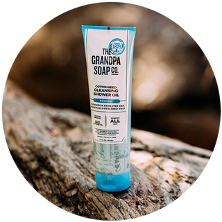 A tube of The Grandpa Soap Co. Cotton Seed Cleansing Shower Oil stands upright on a textured tree branch with a blurred natural background.
