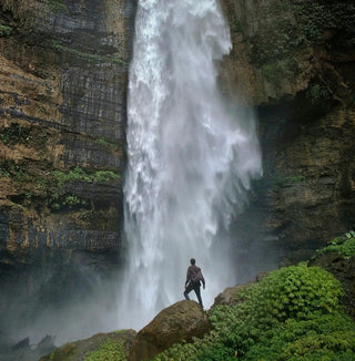A person stands on a rock at the base of a powerful waterfall, surrounded by lush green foliage and high cliffs, gazing up at the rushing water.