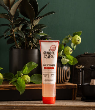 A tube of The Grandpa Soap Co. Apple Cider Vinegar Body Wash stands upright on a wooden surface, surrounded by potted green plants, apples, and grooming accessories against a dark green background.
