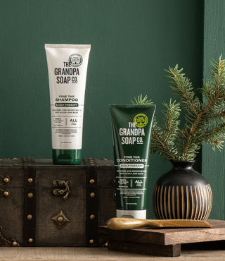 Two tubes of The Grandpa Soap Co. Pine Tar Shampoo and Conditioner are displayed on a wooden chest next to a small vase with pine branches, set against a green wall.