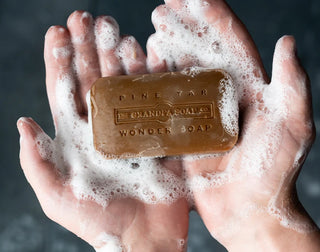A person holds a lathered bar of brown soap labeled Pine Tar, The Grandpa Soap Co., Wonder Soap in their soapy hands.