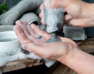 A person dispenses a gray, foamy cleanser onto their hand, with soap suds covering their hands. A towel and a bowl of water are visible on a wooden surface in the background.