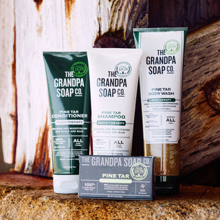 The Pine Tar Collection Bundle by Grandpa Soap Co.—including conditioner, shampoo, body wash, and bar soap—is displayed on a rustic wooden surface with a wooden backdrop. Each product features green, white, and black labels.