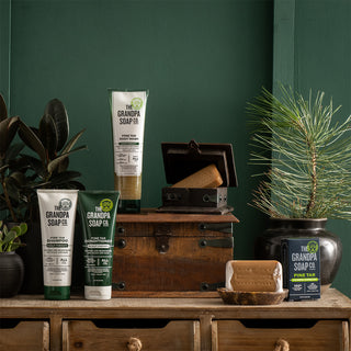 The Grandpa Soap Co. Pine Tar Collection Bundle, featuring shampoo, conditioner, body wash, and bar soap, is displayed on a rustic wooden chest with lush plants in the background.