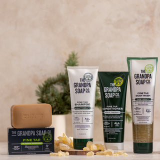 The Pine Tar Collection Bundle from Grandpa Soap Co., featuring bar soap, shampoo, conditioner, and body wash, is displayed on a beige surface with tree resin pieces and a small pine tree in the background.