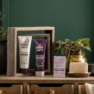 The Witch Hazel Collection Bundle is displayed in a wooden box frame with bottles, a bar soap, its packaging, a soap dish, and a potted plant on a wooden dresser against a green wall.