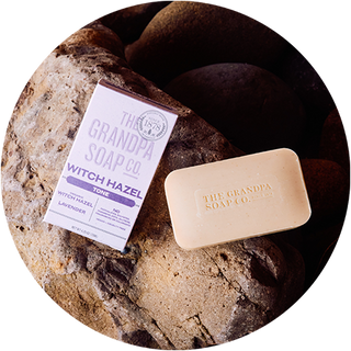 A bar of The Grandpa Soap Co. soap and its box labeled Witch Hazel rest on large, smooth rocks. The soap is beige and the box mentions witch hazel and lavender.